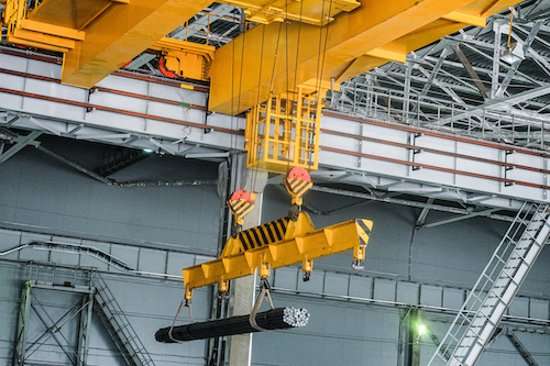 Yellow overhead crane carries cargo in engineering plant shop. Jib crab trolley with hooks and linear traverse.