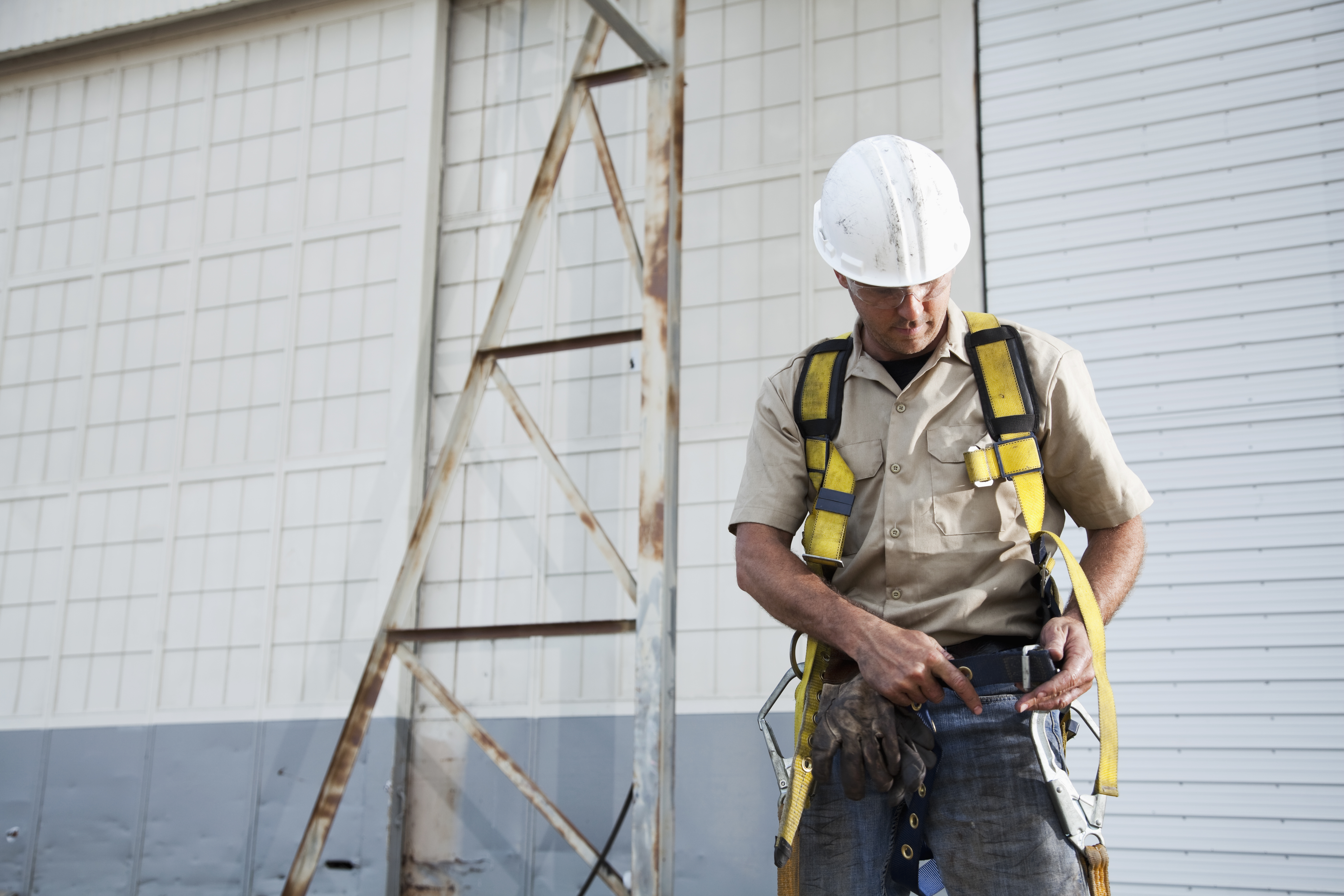 "Worker (30s) with work gloves, hard hat, safety glasses, putting on harness." working at heights training. fall protection equipment