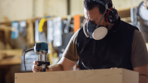 A man wearing a respirator mask and earmuffs uses a router on a piece of wood in a workshop.