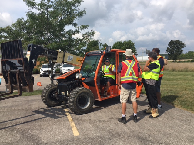 A man in a high visibility vest sitting in a telehandler lift truck demonstrating safe operation to a group of people