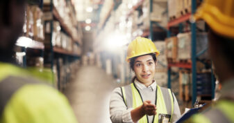 Woman supervisor with clipboard in meeting with other staff, conducting a health and safety meeting in a warehouse.