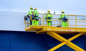 Workers on a yellow self-propelled scissor lift aerial work platform against a blue sandwich panel wall background