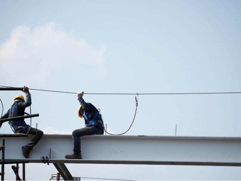Two construction workers working at heights, wearing hard hats and safety harnesses, sit on a large steel beam against a blue sky