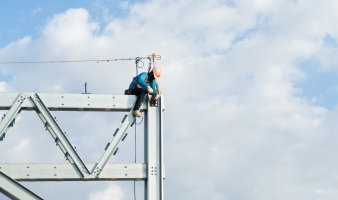 A construction worker wearing a hard hat and safety harness works on a steel beam against a cloudy sky.