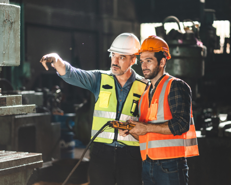 Two workers wearing hard hats and safety vests are in a factory setting. One worker points towards a machine while the other holds a remote control.