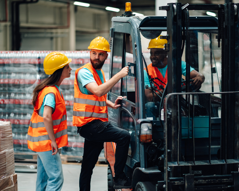 Two people in yellow hard hats and orange safety vests are being trained on a forklift. The Forklift trainer is standing on the ground, and the other is climbing into the forklift.