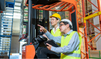 Two people wearing safety vests and hard hats are in a forklift in a warehouse, with one pointing and holding a tablet.