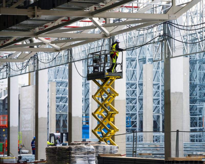 A worker, on a scissor lift, installs equipment on a metal structure inside a large warehouse with blue-tinted glass panels and steel beams.