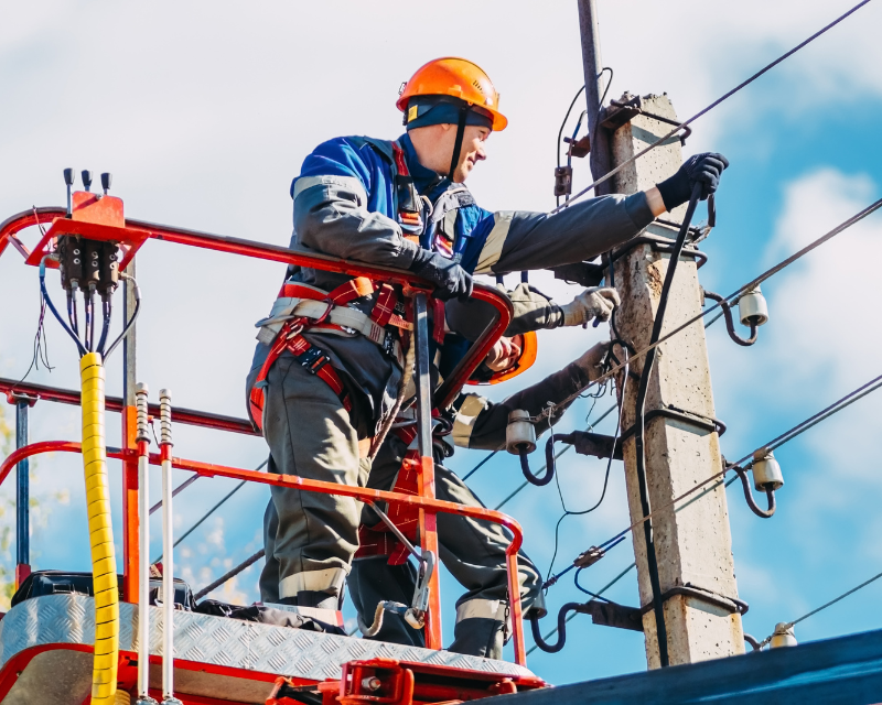 A worker in an orange hard hat and harness on a mobile elevating work platform works on an electrical utility pole, fixing wires.